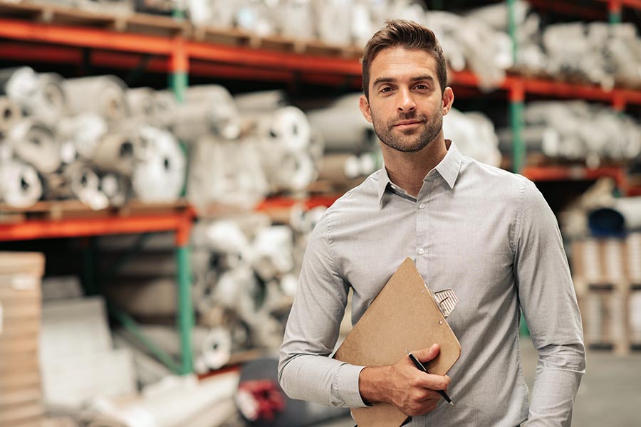 Business Insurance - Business Owner Stands in His Warehouse Holding a Clipboard, Product Filling the Shelves Behind Him