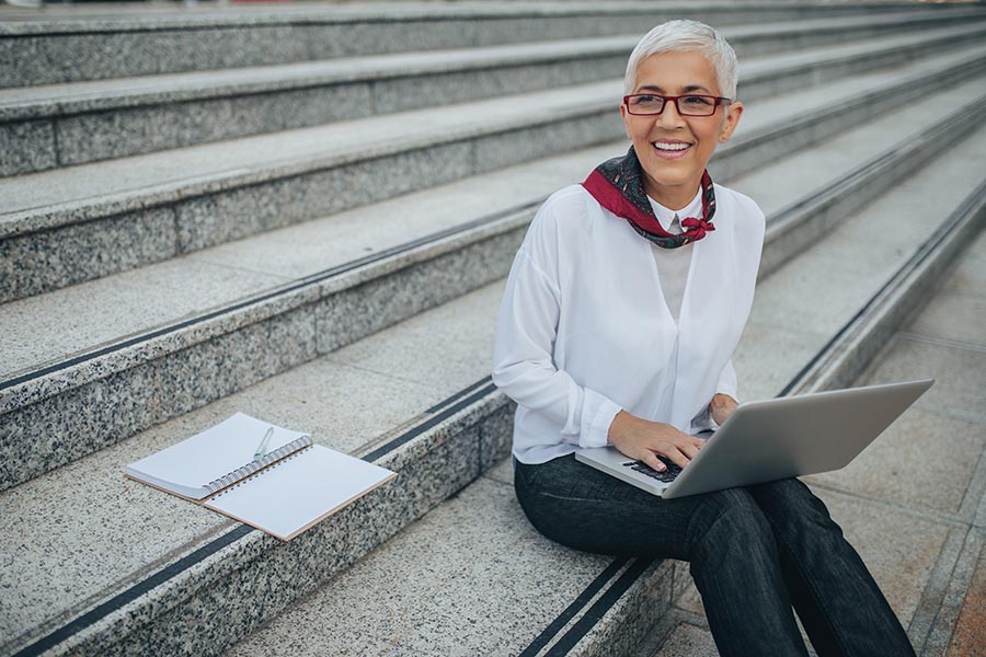 Client Center - Businesswoman Smiles and Sits on the Granite Steps of a Building, a Notebook by Her Side and Computer on Her Lap